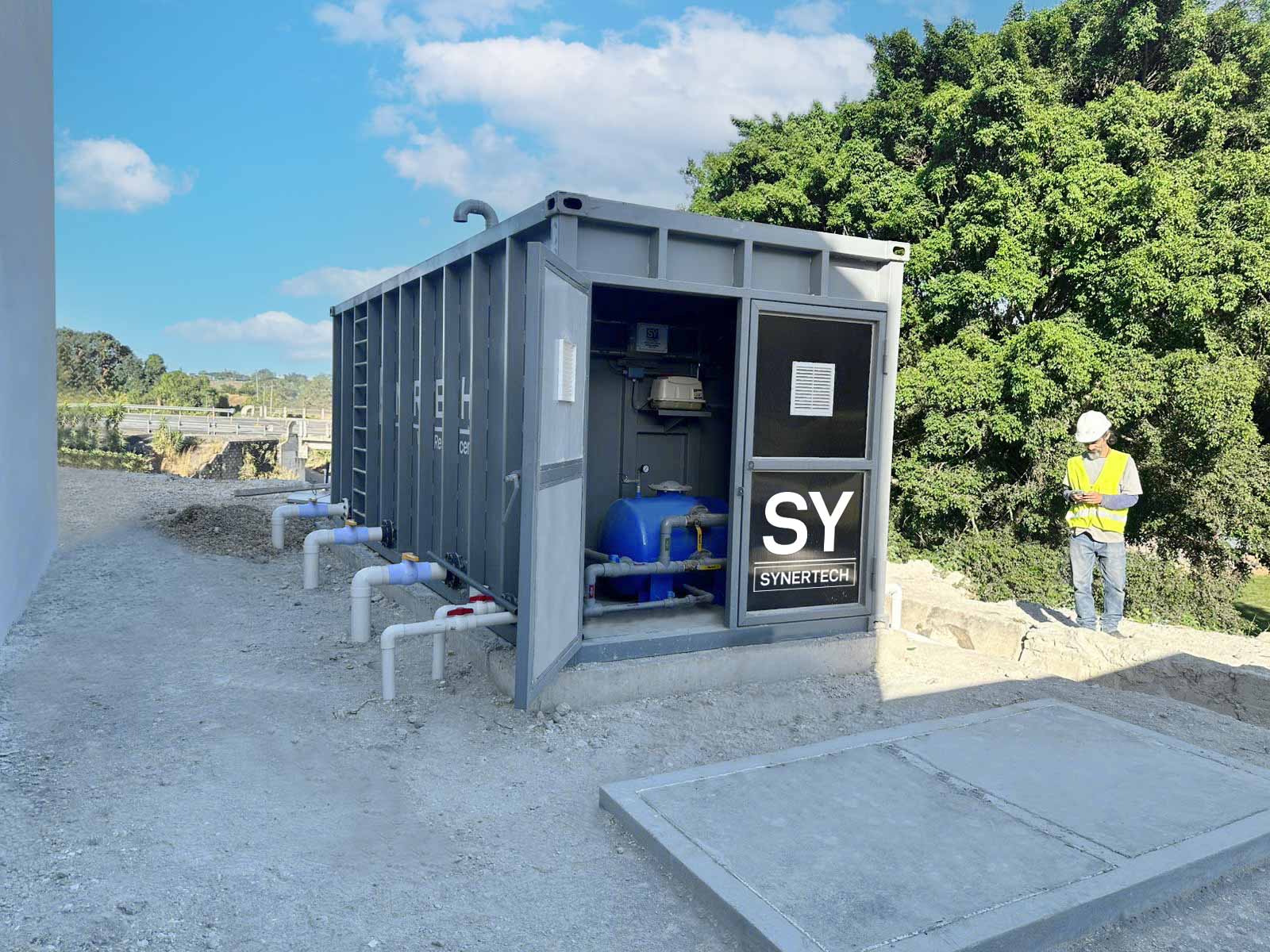 Técnico inspeccionando una planta de tratamiento de aguas residuales modular de color verde marca BIOMAX SYNERTECH, que incluye módulos de pretratamiento, decantación, DAF y oxidación avanzada. La planta está ubicada en un entorno industrial con techo alto y tuberías visibles. - ver más información